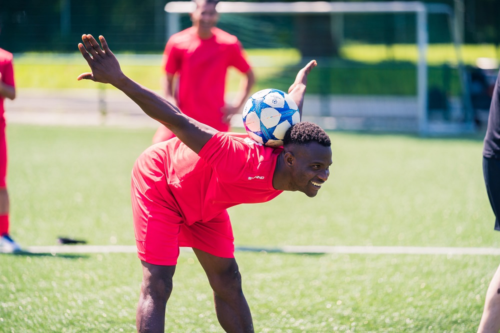 Ein junger Mann in roter Sportbekleidung balanciert einen Fußball auf dem Kopf und streckt dabei einen Arm aus, während er lächelt. Im Hintergrund sind weitere Personen und ein Fußballfeld zu sehen.