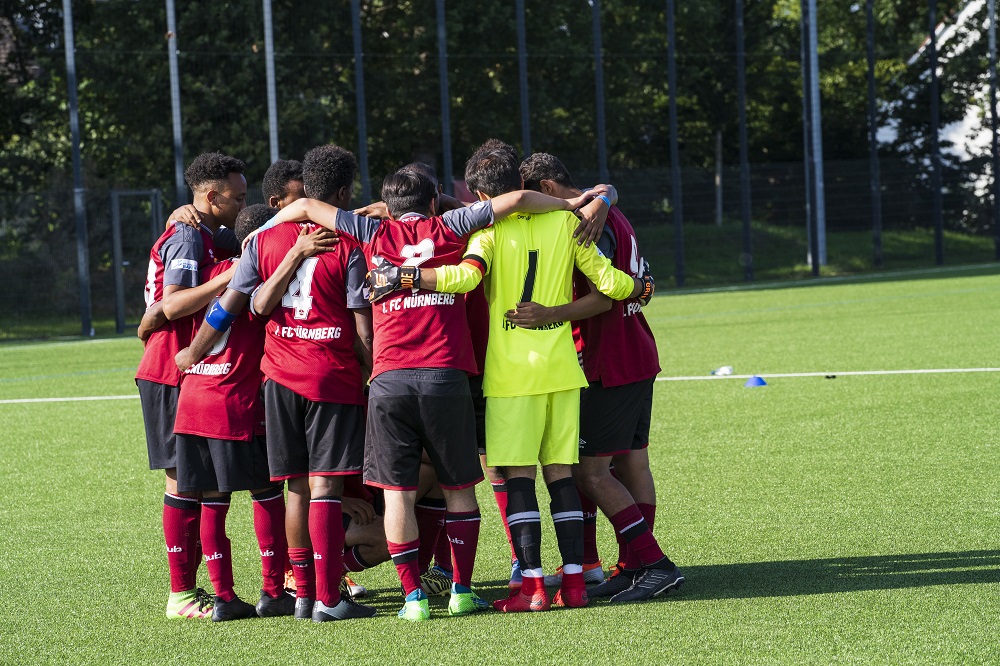 Fußballmannschaft im Kreis, Spieler mit roten Trikots und schwarzer Hose stehen auf einem grünen Rasenplatz eng zusammen und bilden einen Kreis mit verschlungenen Armen.