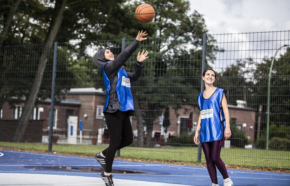 Zwei Frauen mit blauen Westen stehen auf einem Basketballfeld und spielen. Die eine wirft einen Ball hoch, die andere blickt ihr dabei zu. Im Hintergrund sind Bäume und ein Zaun zu sehen.