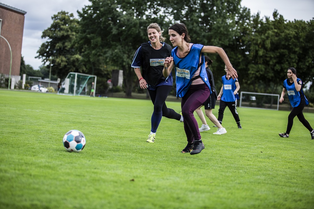 Frauenfußballspiel im Rahmen des "Willkommen im Fußball"-Ladies Cups mit Spielerinnen in blauem und weißem Trikot auf einem grünen Rasenfeld. Im Hintergrund sind weitere Spielerinnen und Bäume zu sehen.