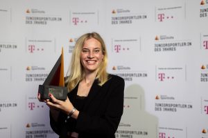 Hannah Peters, eine junge Frau mit blonden Haaren, lächelt in die Kamera und hält eine gläserne Trophäe in den Händen. Im Hintergrund ist ein Banner mit dem Aufdruck "Juniorsportler des Jahres" zu sehen.