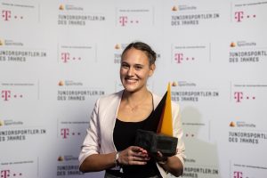 Stabhochspringerin Sarah Vogel hält lächelnd die Auszeichnung "Juniorsportlerin des Jahres" entgegen. Im Hintergrund ein Banner mit dem Logo des Sponsors und dem Titel der Auszeichnung.