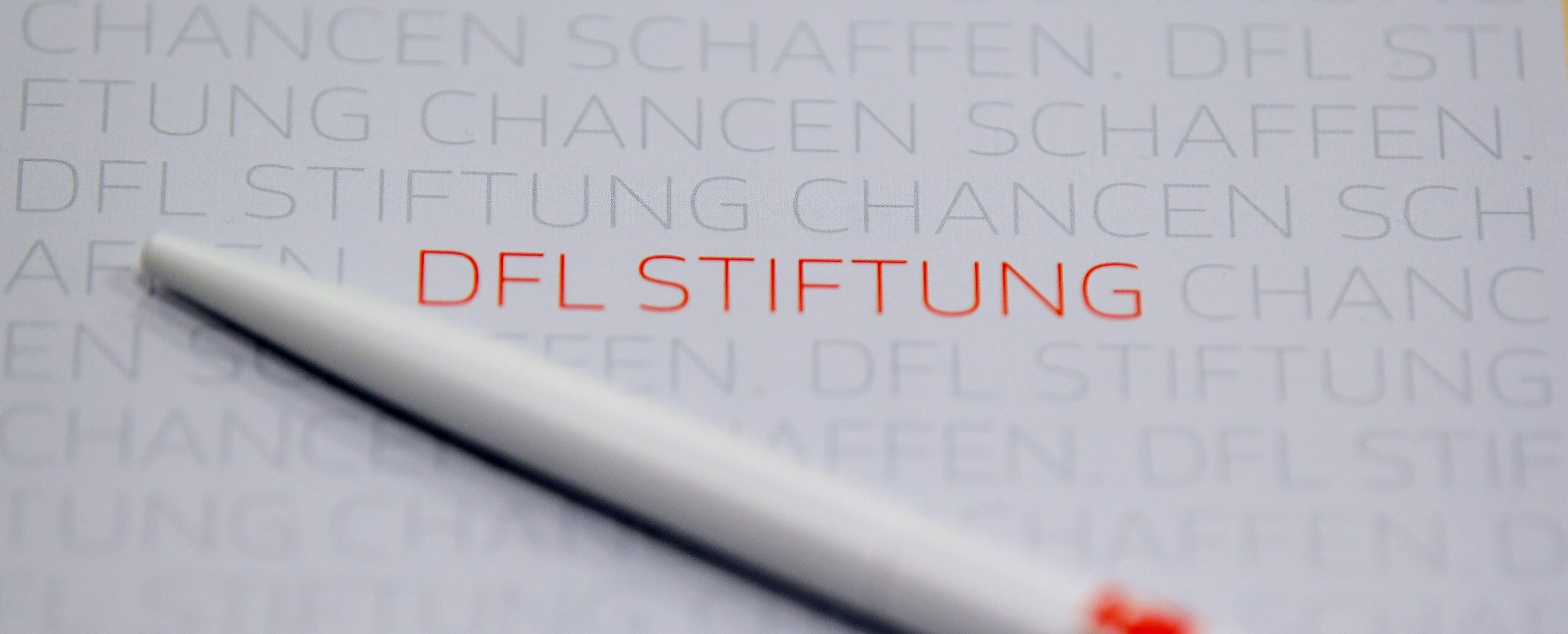 DFL Stiftung - Nahaufnahme eines Stifts, der über ein transparentes Blatt mit dem wiederholten Schriftzug "DFL STIFTUNG Chancen schaffen" fährt.