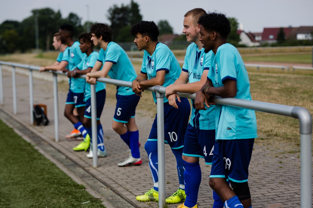 Fußballspieler in blauen Trikots lehnen an einer Absperrung am Spielfeldrand, während des regionalen "Willkommen im Fußball"-Cups in Wolfsburg.