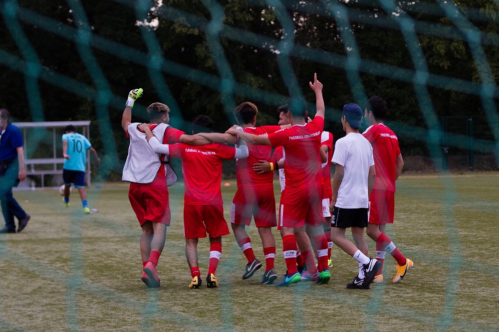 Eine Gruppe von jungen Fußballspielern im roten Trikot feiert gemeinsam mit erhobenen Armen auf einem grünen Fußballfeld. Im Hintergrund ist ein grünes Absperrgitter zu sehen.