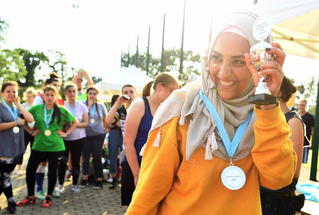 Lächelnde Frau mit Kopftuch hält einen Pokal in die Höhe, umgeben von Teilnehmenden eines Sportevents. Im Hintergrund sind weitere Menschen und Bäume zu sehen.
