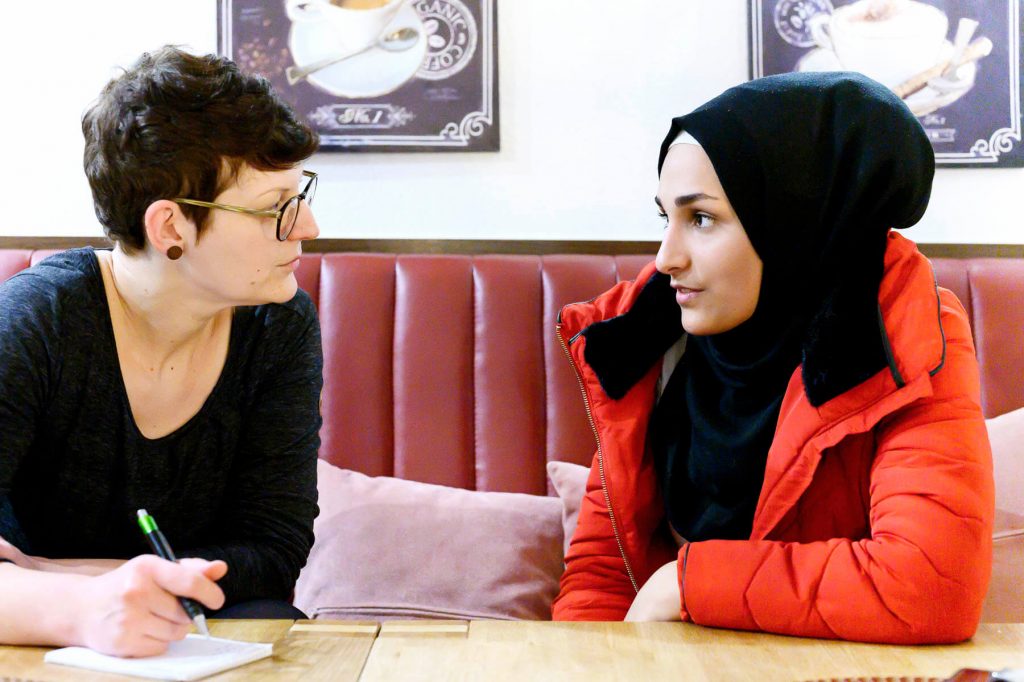 Zwei junge Frauen sitzen sich an einem Holztisch gegenüber und unterhalten sich. Die Frau links schreibt Notizen, während die Frau rechts, mit Kopftuch, ihr aufmerksam zuhört. Im Hintergrund ist ein rotes Polster an der Wand zu sehen.