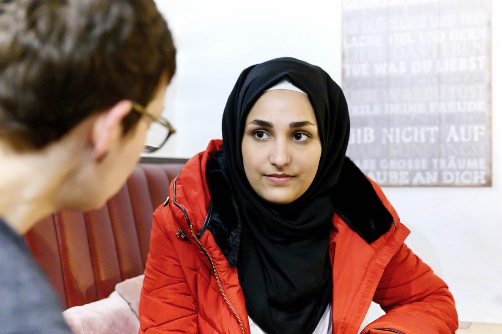 Eine Frau mit Kopftuch und roter Jacke sitzt einer anderen Person gegenüber und blickt in die Kamera. Im Hintergrund ist ein Plakat mit der Aufschrift "Gib nicht auf. Deine Träume rühren dich".