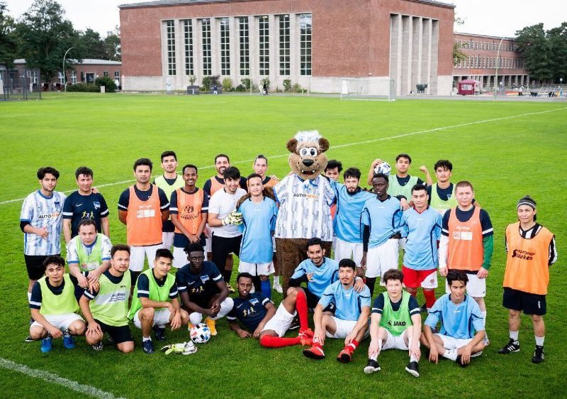 Zwei Jugendliche spielen im Freien Fußball. Links ein Spieler im blauen Trikot mit dem Schalker Logo und dem Schriftzug "Gazprom", rechts ein Spieler in rotem Trikot mit dem Eintracht Frankfurt Logo. Im Hintergrund sind weitere spielende Kinder erkennbar.