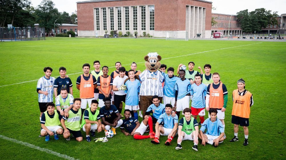 Zwei Jugendliche spielen im Freien Fußball. Links ein Spieler im blauen Trikot mit dem Schalker Logo und dem Schriftzug "Gazprom", rechts ein Spieler in rotem Trikot mit dem Eintracht Frankfurt Logo. Im Hintergrund sind weitere spielende Kinder erkennbar.