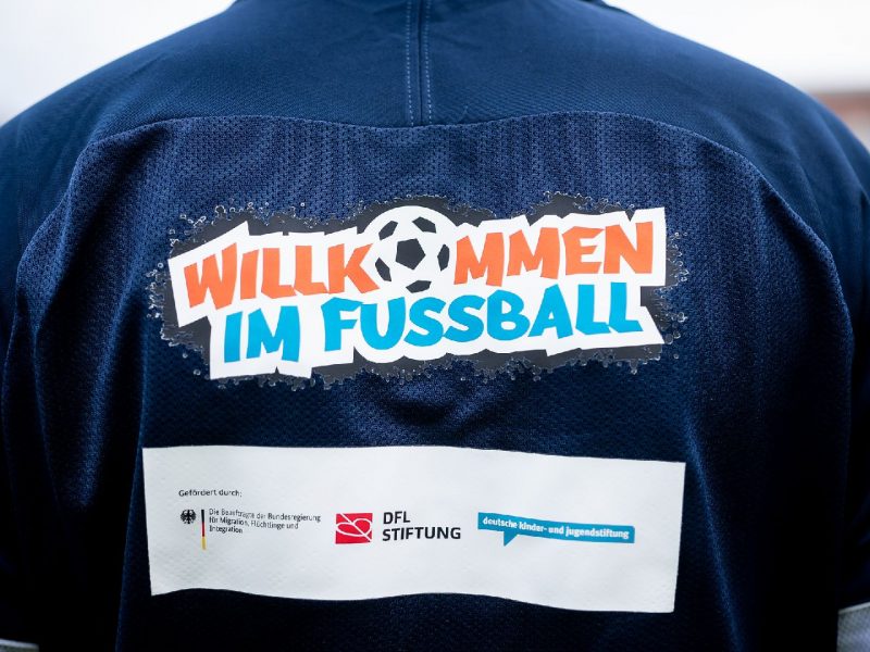 Eine Frau mit rotem Schal, ein Mann im Anzug und eine weitere Frau stehen in einer Sporthalle und unterhalten sich. Ein Jugendlicher im roten Trainingsshirt hält einen Fußball in die Hand.