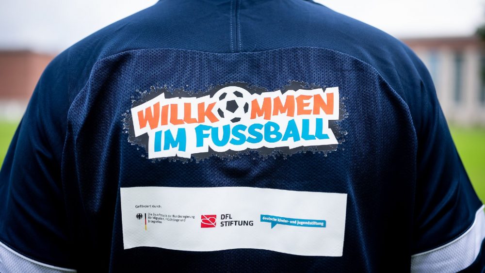 Eine Frau mit rotem Schal, ein Mann im Anzug und eine weitere Frau stehen in einer Sporthalle und unterhalten sich. Ein Jugendlicher im roten Trainingsshirt hält einen Fußball in die Hand.