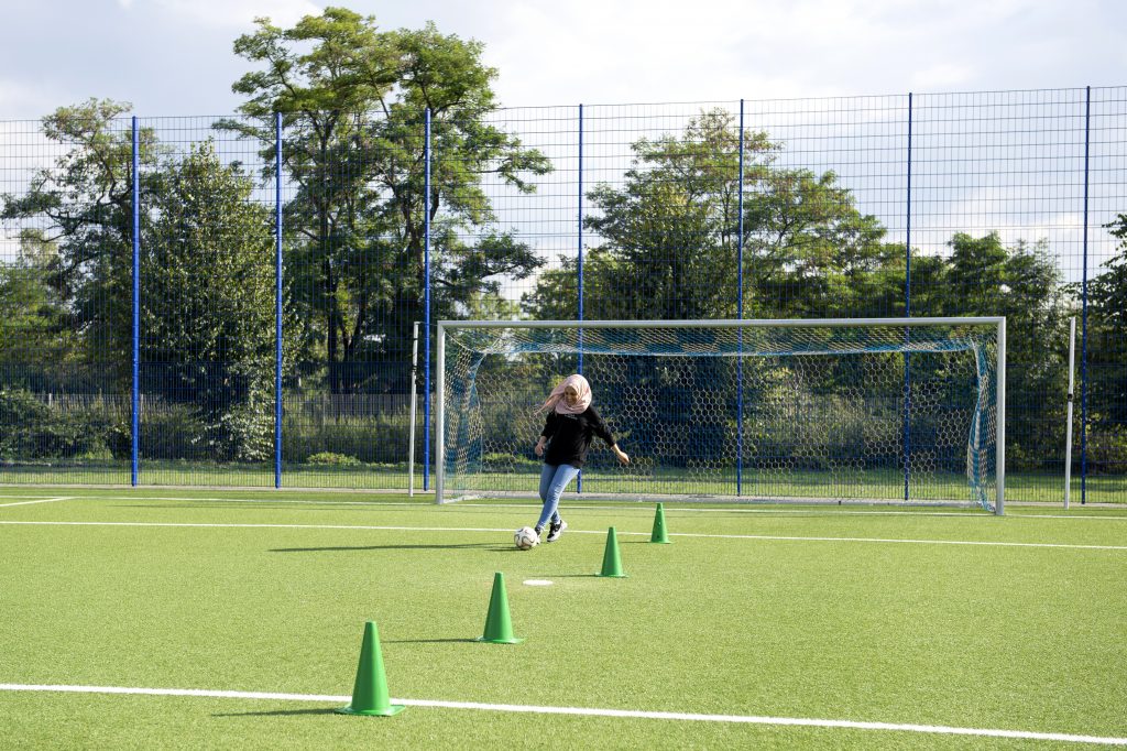 Ein junges Mädchen dribbelt einen Fußball geschickt um Hütchen auf einem grünen Kunstrasenplatz. Im Hintergrund steht ein Fußballtor und ein Maschendrahtzaun.
