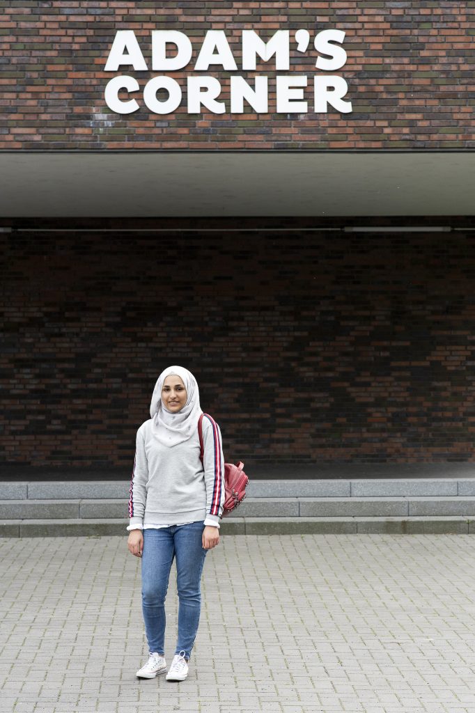 Eine junge Frau mit Kopftuch steht vor einer roten Backsteinwand mit der Aufschrift "ADAM'S CORNER". Sie trägt einen grauen Hoodie, Jeans und weiße Sneaker und hat einen Rucksack über der Schulter.