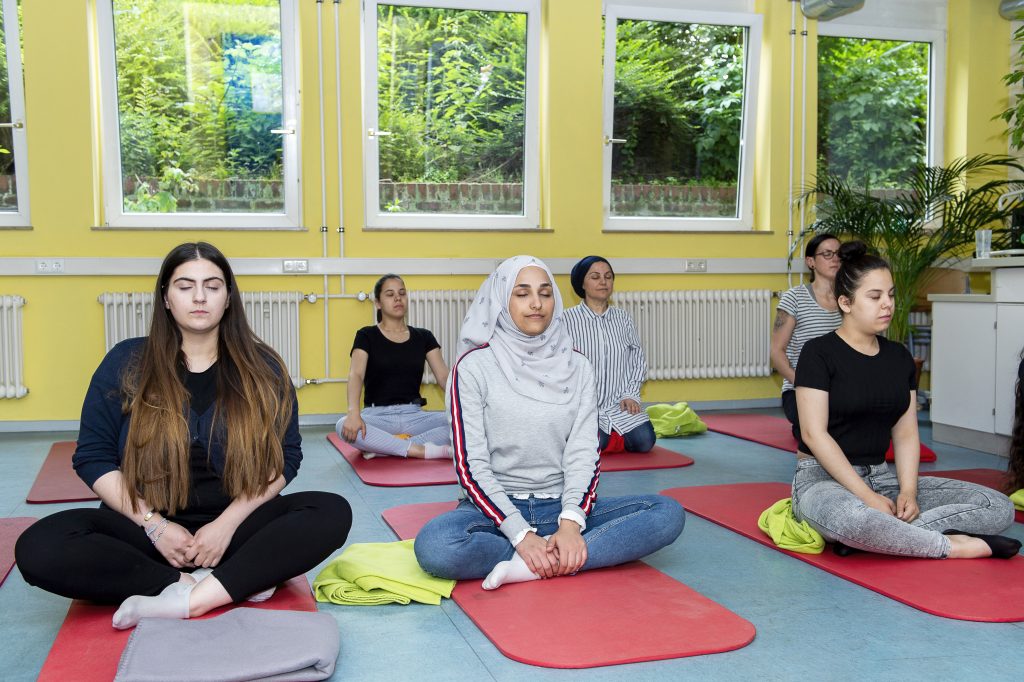 Eine Gruppe junger Frauen sitzt in einem hellen Raum auf Yogamatten im Schneidersitz und scheint an einer Entspannungsübung teilzunehmen. Einige tragen ein Kopftuch. Im Hintergrund sind Heizkörper und Fenster zu sehen.