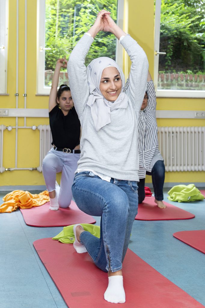 Eine junge Frau mit Kopftuch und Jeans kniet auf einer roten Matte und führt eine Dehnübung durch. Sie lächelt in die Kamera. Im Hintergrund sind weitere Personen bei ähnlichen Übungen zu sehen.