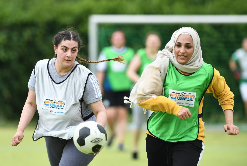 Zwei junge Frauen mit Kopftuch und Sportkleidung spielen Fußball im Freien. Die Frau links fokussiert den Ball, die Frau rechts läuft neben ihr her. Im Hintergrund sind weitere Spielerinnen und ein Fußballtor zu sehen.