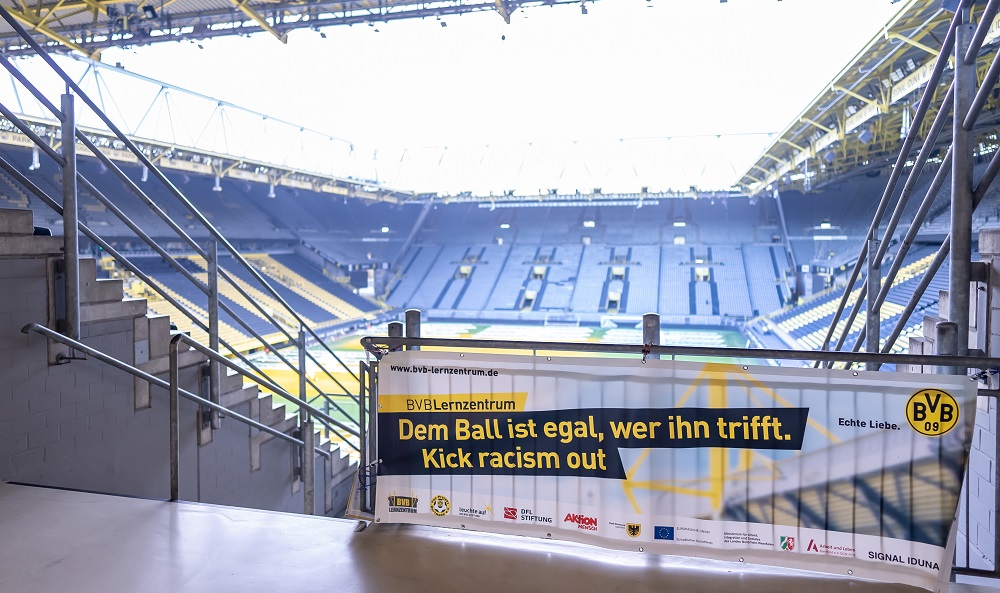 Eine Gruppe von Jugendlichen, gemischt aus deutschen und kenianischen Fußballspielern, posiert auf einem Fußballfeld für ein Gruppenfoto. Sie tragen alle weiße und blaue Sportkleidung. Im Hintergrund befindet sich ein Gebäude und ein Fußballtor.