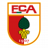 Logo FC Augsburg Eine Gruppe von Kindern und Jugendlichen sitzt auf dem Fußballrasen vor farbenfrohen, bogenförmigen Pappaufstellern. Im Hintergrund ist ein volles Stadion zu sehen.