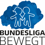 Ein Mann mit dunklen Haaren lächelt und gestikuliert, während er mit einer anderen Person im Hintergrund spricht. Beide tragen lila T-Shirts mit der Aufschrift "SCHÖN DASS DU DABIST".