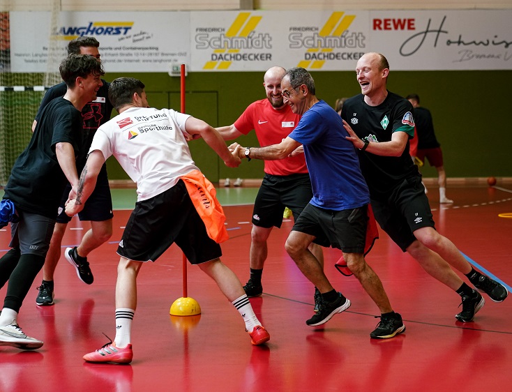 Eine Gruppe von Kindern und Jugendlichen sitzt im Kreis auf dem Boden einer Sporthalle. Ein Mann sitzt mit ihnen im Kreis und hält einen Fußball. Die Kinder sind unterschiedlich gekleidet und tragen verschiedene Westen. Im Hintergrund sind die Holzwände der Halle und eine Anzeigetafel zu sehen.