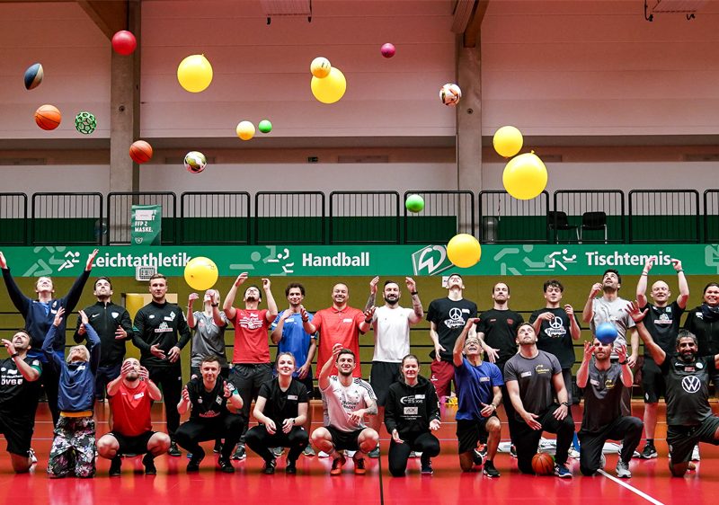 Ein junger Fußballspieler im roten Trikot dribbelt in einer Sporthalle an einem Gegenspieler vorbei. Im Hintergrund sind weitere Jugendliche beim Fußballspielen zu sehen.