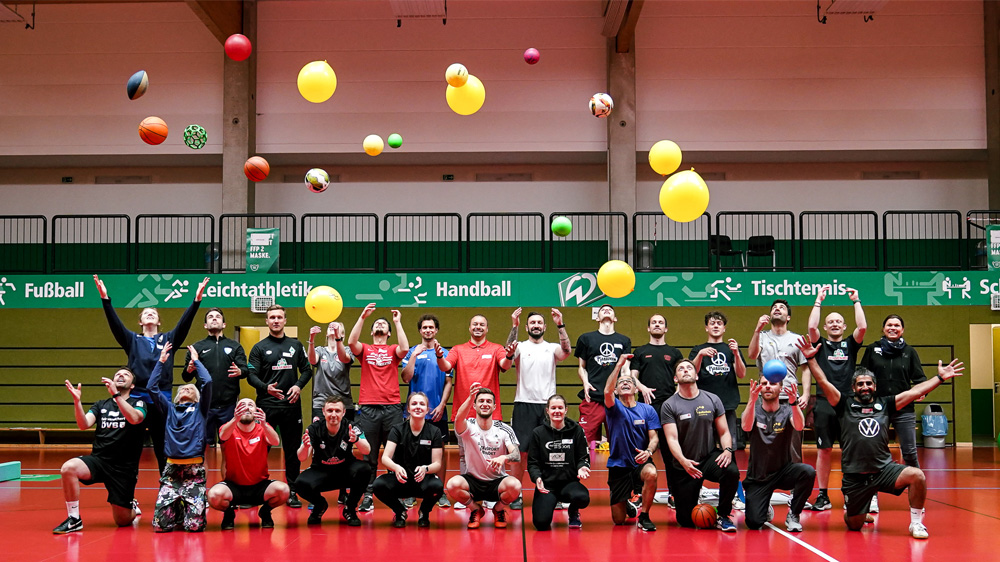 Ein junger Fußballspieler im roten Trikot dribbelt in einer Sporthalle an einem Gegenspieler vorbei. Im Hintergrund sind weitere Jugendliche beim Fußballspielen zu sehen.