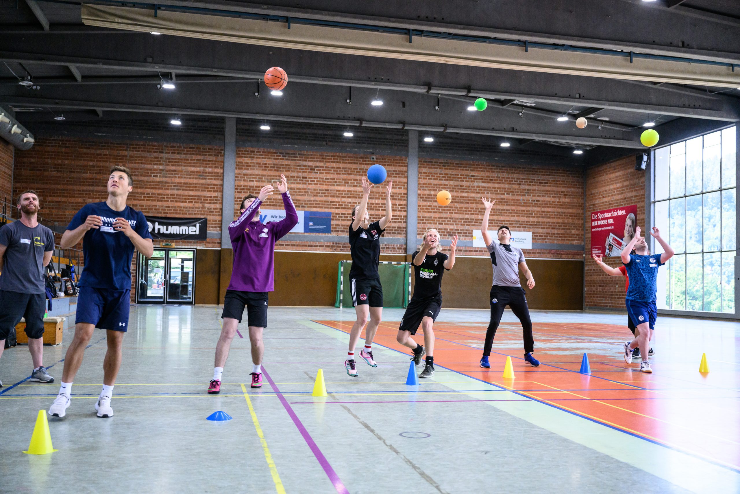Rasengras auf einem Fußballfeld, mit hoch fliegenden Grashalmen. Im Hintergrund ein verschwommener roter Bereich, vermutlich die Sitztribüne eines Stadions.