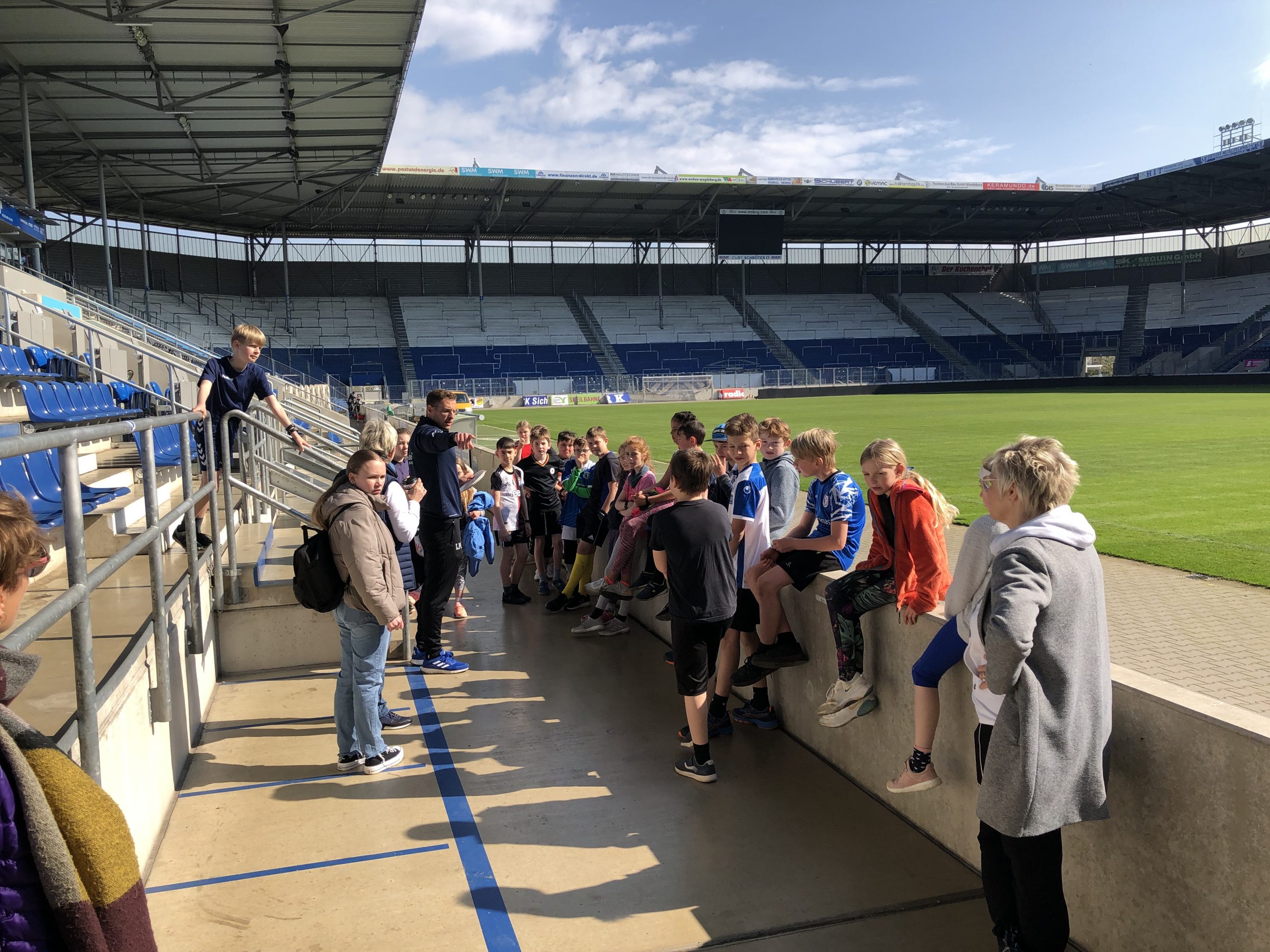 Glückliche Schülerinnen und Schüler der Michael-Ende-Schule aus Frankfurt am Main halten gemeinsam den Siegerpokal des U10-Turniers des Projekts "Fußball trifft Kultur" hoch. Ein Mann mit Hemd und Jeans gibt Daumen hoch. Im Hintergrund sind weitere Menschen und ein Fußballfeld zu sehen.