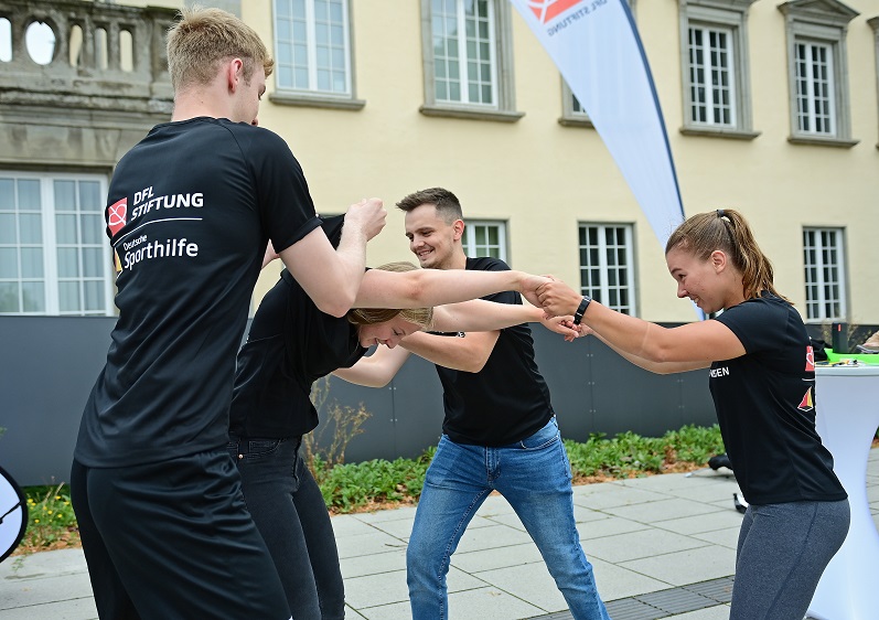 Gruppe von sieben Personen steht auf einer Bühne vor einem Hintergrund mit dem Logo "ANSTOSS 2019" und dem DFL-Logo. Sie halten Urkunden in den Händen, vermutlich als Anerkennung für ihre Arbeit bei der DFL Stiftung.