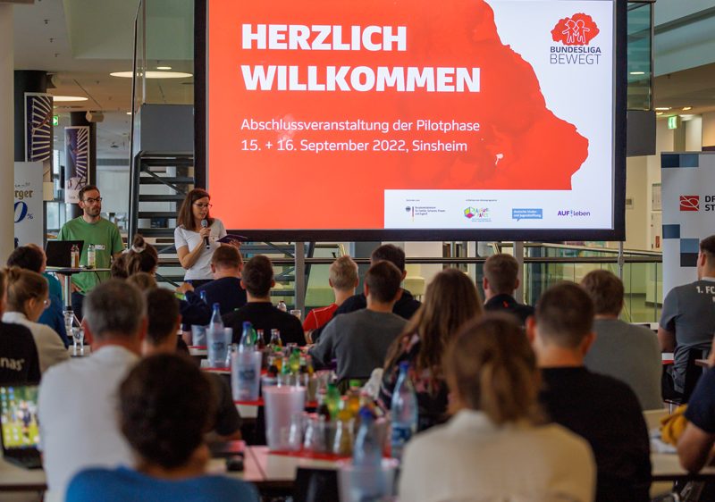 Eine Gruppe von Erwachsenen steht vor einem Banner mit der Aufschrift "Fußball trifft Kultur" und dem Logo der DFL Stiftung. Im Hintergrund ist ein Fußballfeld mit spielenden Kindern erkennbar.