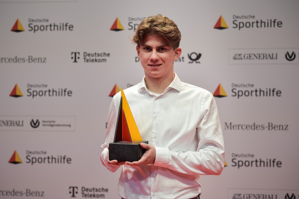 Fußballspieler Davie Selke, porträtiert mit halbseitigem Schatten, rechts im Gesicht strahlend, links im Schatten, mit dem Schriftzug "SEITE AN SEITE" im Bild. Logo der DFL Stiftung und Deutsche Sporthilfe im unteren Bereich.