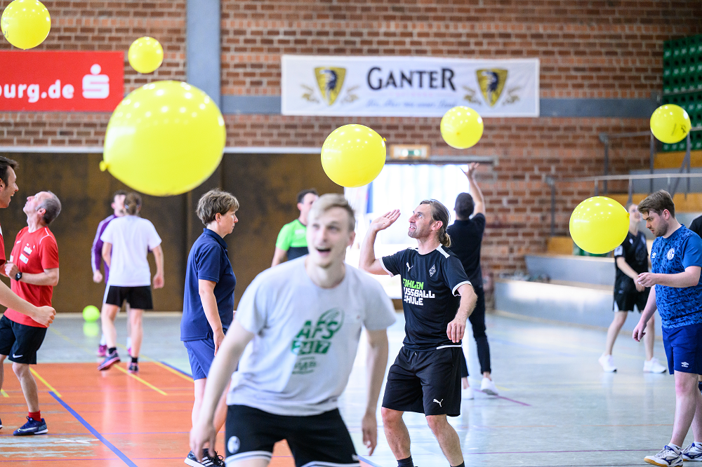 Eine Gruppe von Teilnehmenden steht in einer Sporthalle und spielt mit gelben Bällen. Im Vordergrund ein Mann in Sportkleidung, der einen Ball hochwirft. Im Hintergrund weitere Personen, die ebenfalls aktiv sind. Die Szene zeigt eine Qualifizierungsveranstaltung für Bewegungskoordinator*innen, bei der neue Spielformen erlernt werden.
