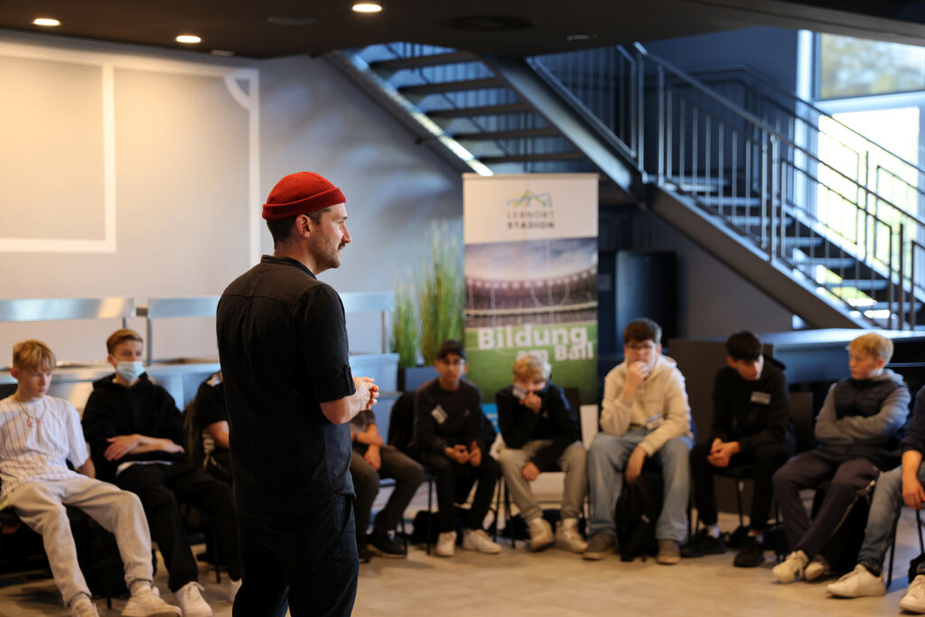 Dominic Ionescu, Koordinator der Stadionschule Bielefeld, hält vor einer Gruppe Jugendlicher einen Vortrag. Im Hintergrund eine Treppe und ein Banner mit der Aufschrift "Bildung im Blick".