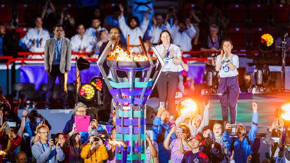 Entzündung der Flamme der Nationalen Sommerspiele der Special Olympics 2023 bei der Eröffnungsfeier. Ein Mensch entzündet eine abstrakt gestaltete Fackel, umgeben von weiteren Personen und jubelnden Zuschauern. Im Hintergrund ist ein blau beleuchtetes Stadion zu sehen.