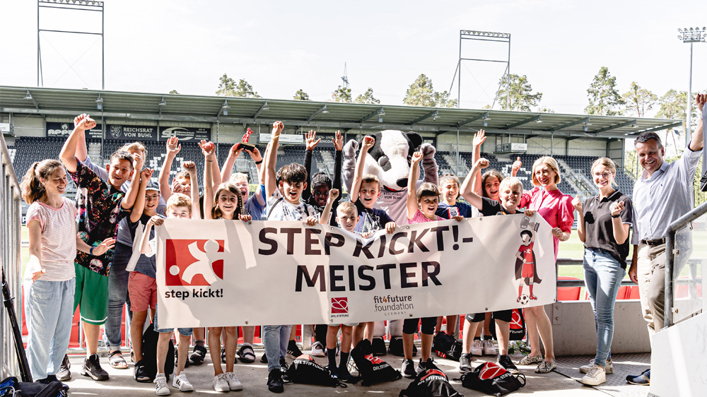 Eine Gruppe von Kindern und Jugendlichen steht strahlend mit hoch erhobenen Armen vor einer Tribüne. Sie halten ein Banner mit der Aufschrift "STEP KICKT! - MEISTER". Im Hintergrund ist ein Fußballstadion zu sehen.