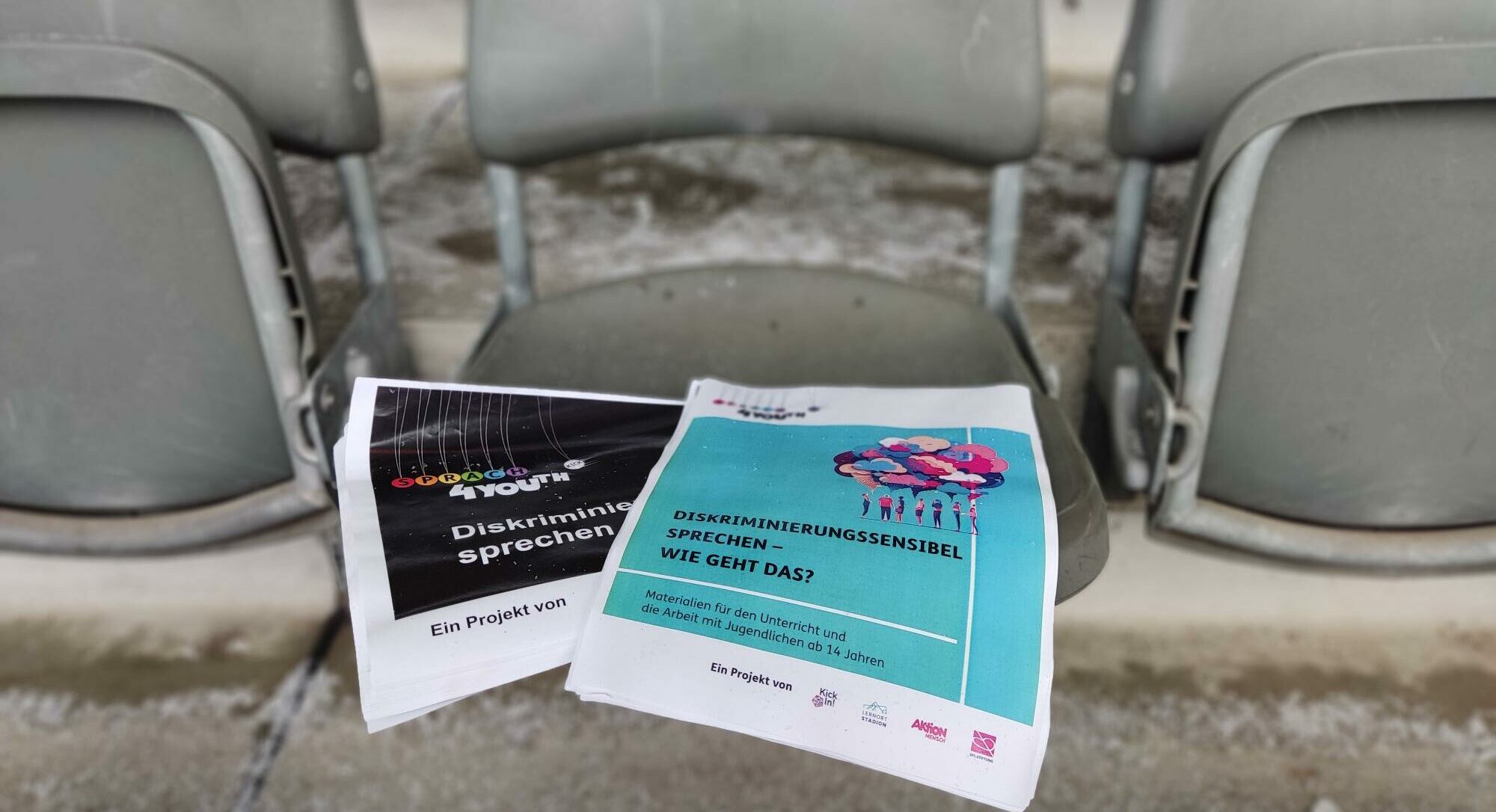 Zwei Flyer liegen auf einem grauen Sessel im Olympiastadion. Der eine Flyer ist schwarz mit dem Aufdruck "Diskriminierung sprechen", der andere blau mit dem Titel "Diskriminierungssensibel – Wie geht das?". Darunter steht "Materialien für die Arbeit mit Jugendlichen ab 14 Jahren". Beide Flyer tragen den Hinweis "Ein Projekt von Kick".