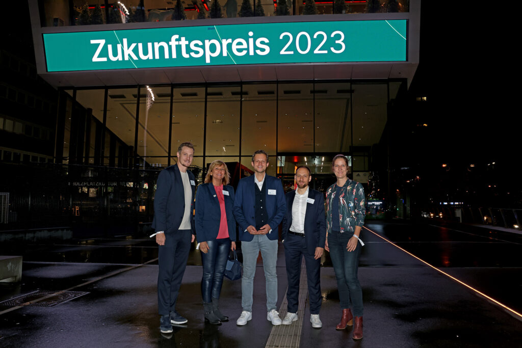 Eine Gruppe von sechs Personen steht vor dem beleuchteten Stadion. Über ihnen ist ein großes digitales Display mit der Aufschrift "Zukunftspreis 2023" zu sehen.