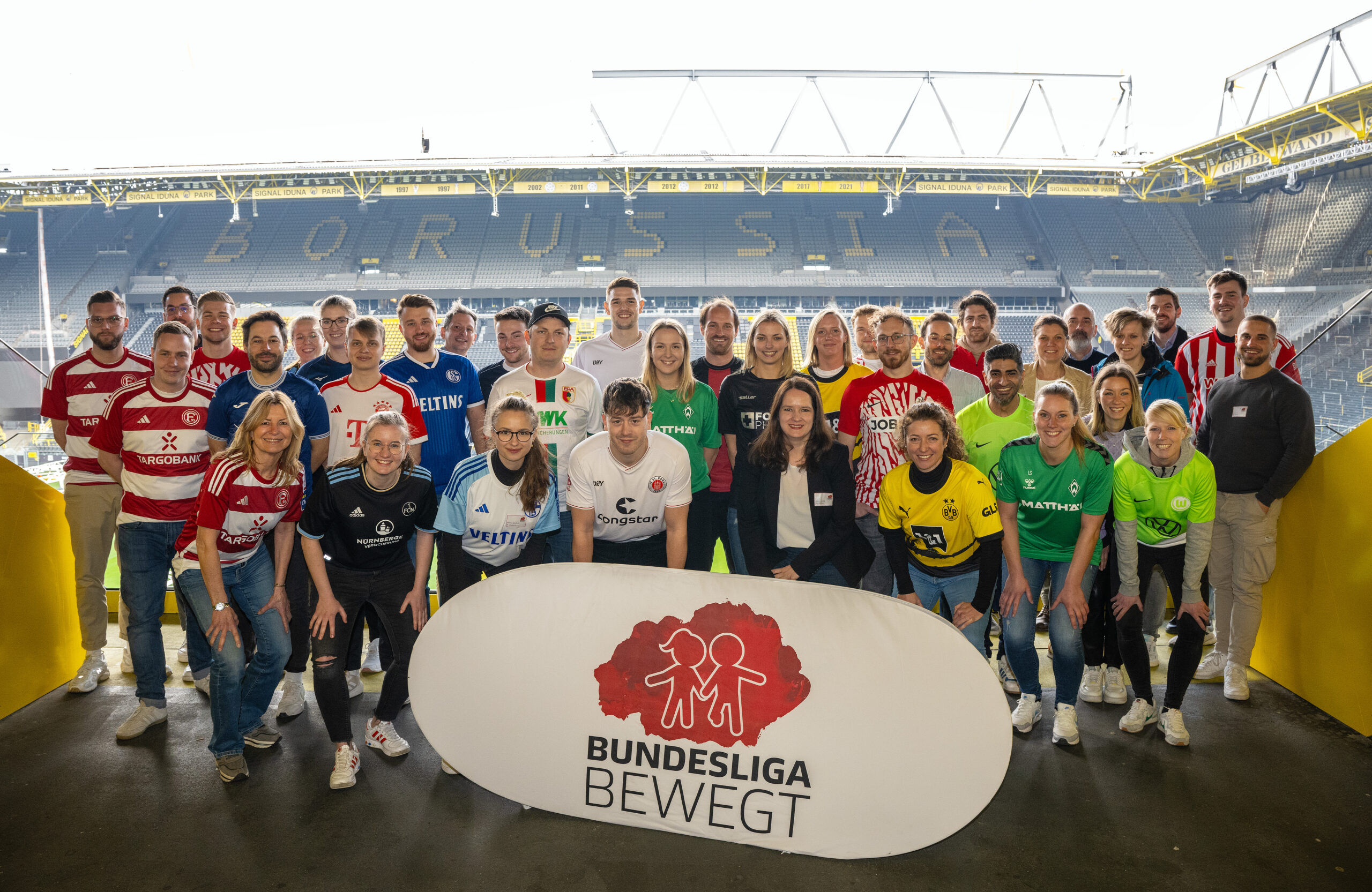 Eine große Gruppe von Menschen steht im Signal Iduna Park in Dortmund vor der Logo-Wand "Bundesliga bewegt" der DFL Stiftung. Viele tragen Trainingsanzüge oder Trikots verschiedener Bundesligavereine. Es handelt sich um ein Netzwerktreffen der DFL Stiftung.