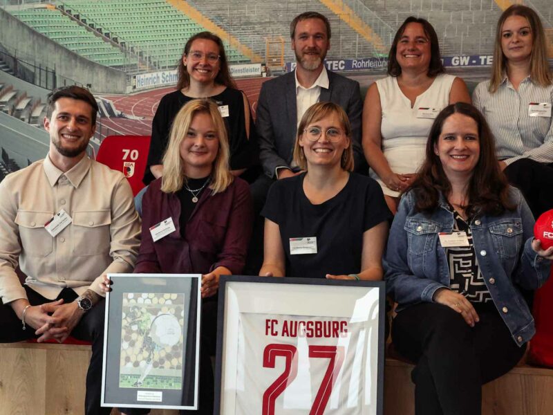 Eine Gruppe von etwa zehn Personen sitzt oder steht lächelnd vor einer Wand mit Stadionmotiven. Im Mittelpunkt hält eine Frau ein gerahmtes Foto, auf dem vermutlich eine Sportanlage zu sehen ist. Alle tragen Kleidung mit dem Logo des FC Augsburg oder Namensschilder, was auf ein gemeinsames Projekt oder eine Veranstaltung im Rahmen der DFL Stiftung hindeutet.
