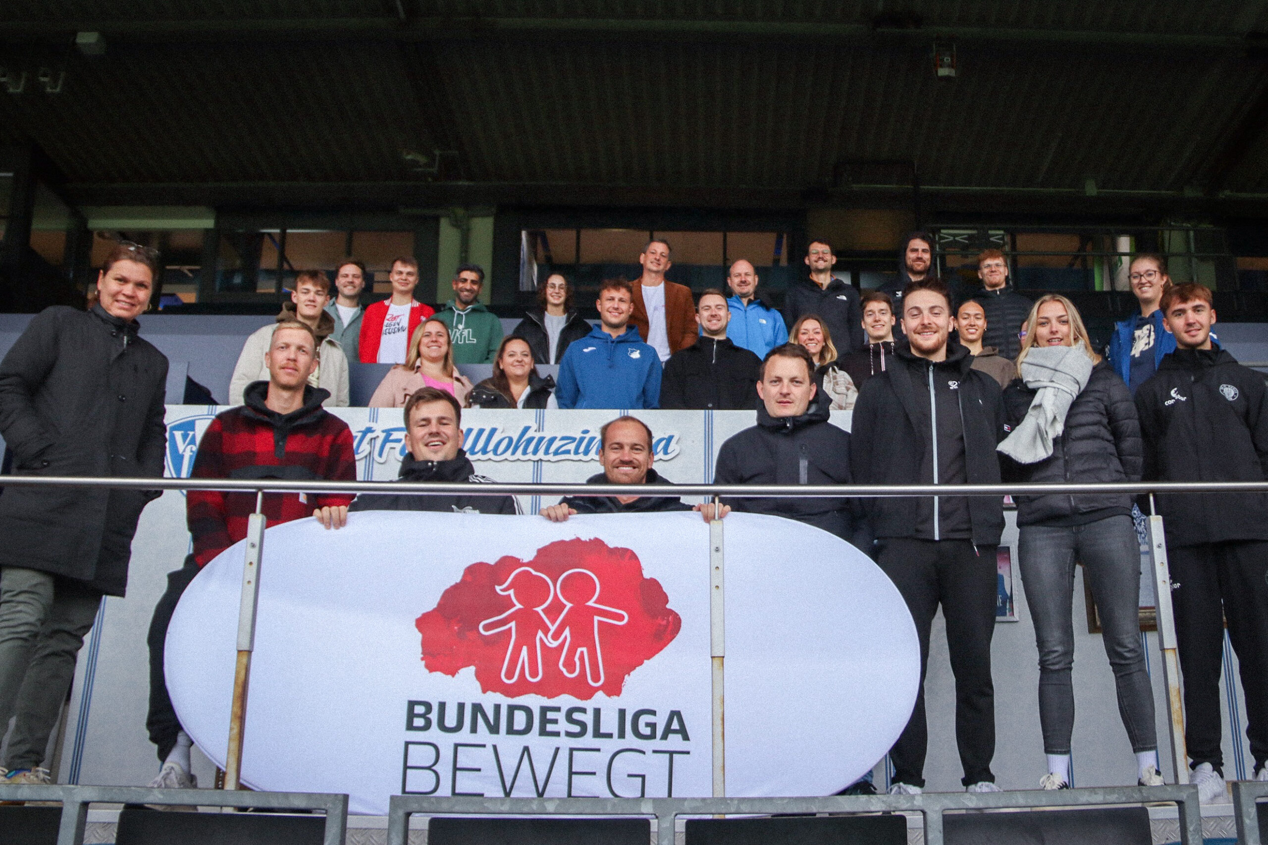 Eine Gruppe von Menschen steht auf einer Tribüne eines Fußballstadions und hält ein Banner mit dem Aufdruck "BUNDESLIGA BEWEGT" und einem roten Symbol, das stilisierte Figuren von Erwachsenen und Kindern darstellt.