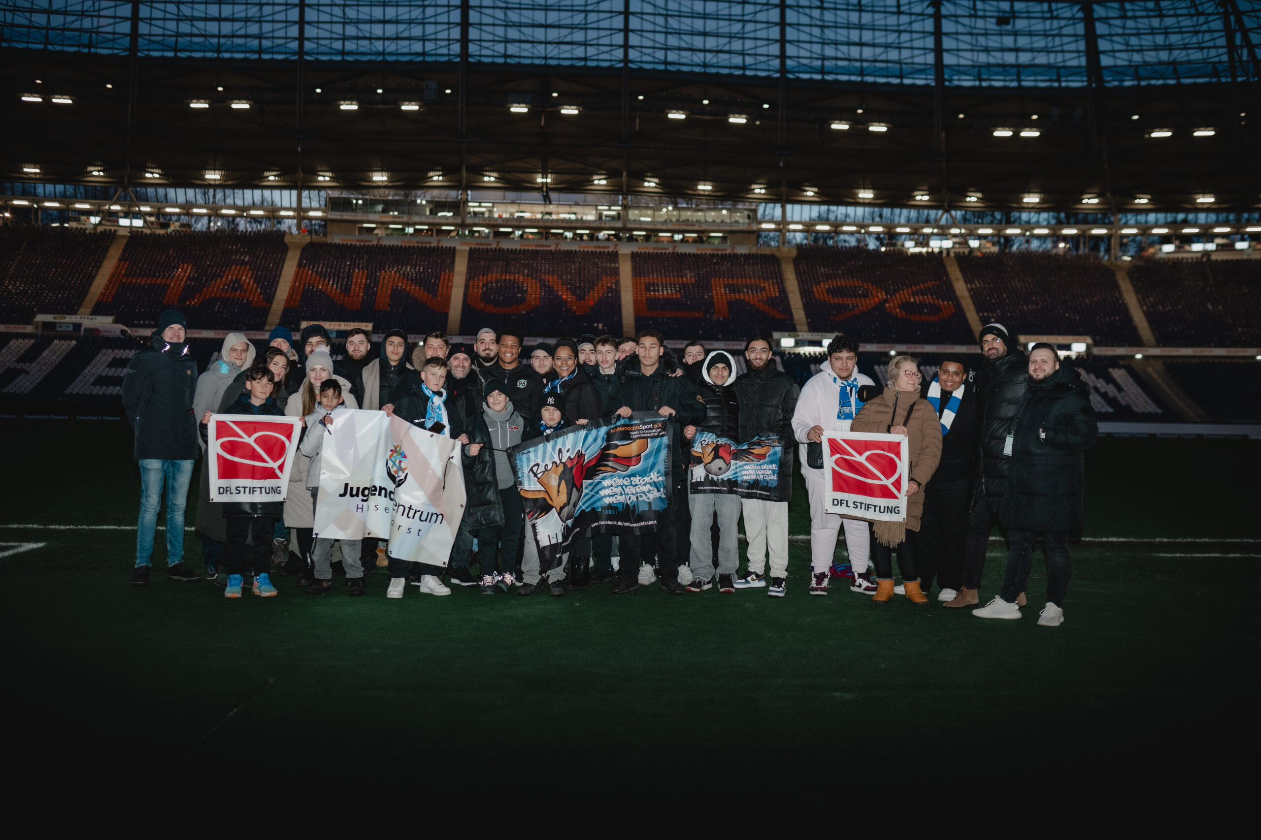 Eine Gruppe von Menschen steht auf dem Rasen eines Fußballstadions. Einige halten Banner mit dem Logo der DFL Stiftung und dem Schriftzug "Jugendzentrum" hoch. Im Hintergrund sind die leeren Zuschauertribünen zu sehen.