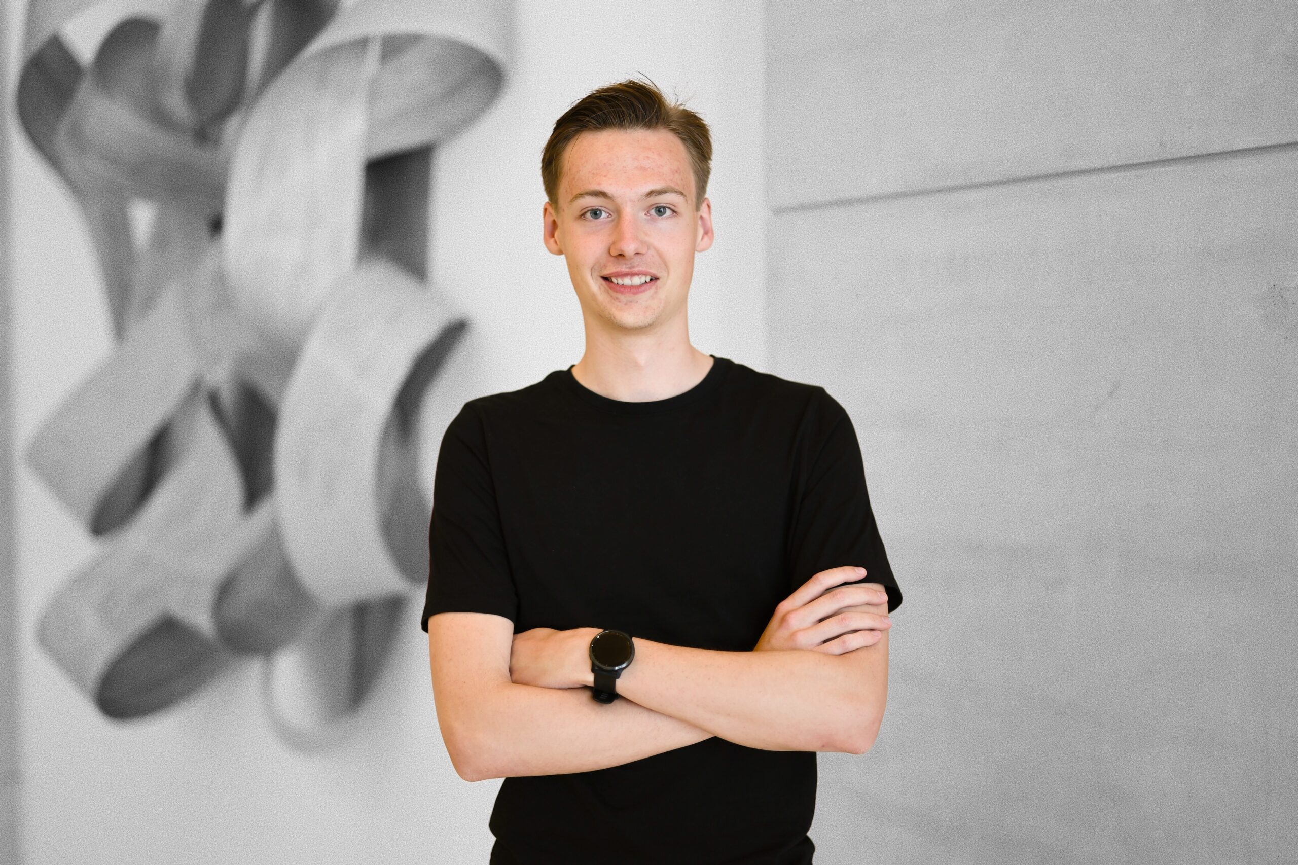 Junge Person mit verschränkten Armen, lächelnd, im Hintergrund eine abstrakte Wandinstallation – Teilnehmer des Treffens des Jugendgremiums der DFL Stiftung.