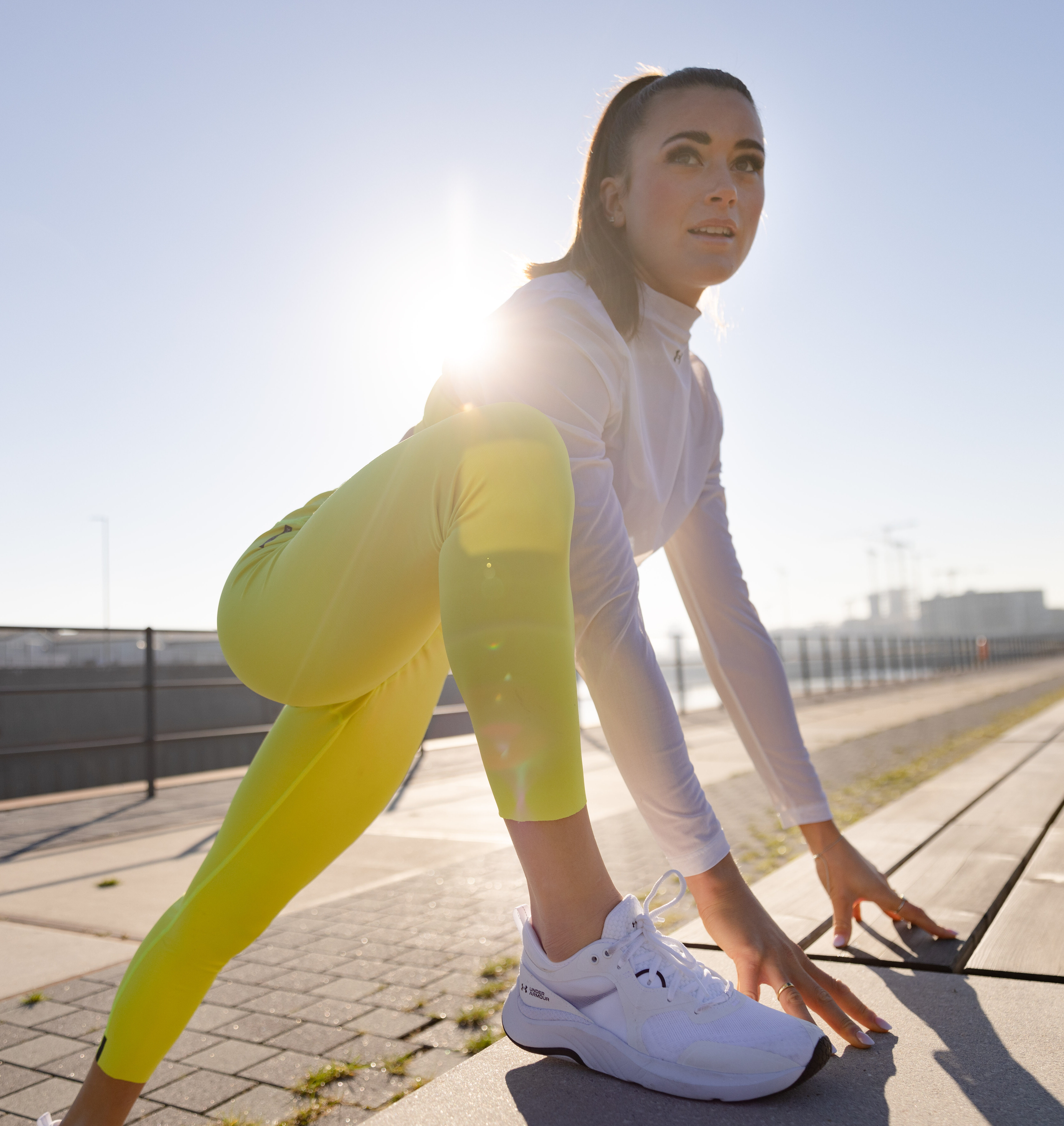 Eine Frau in Sportkleidung macht Dehnübungen auf einer Laufbahn im Freien. Die Sonne scheint hell und betont die dynamische Pose der Sportlerin.