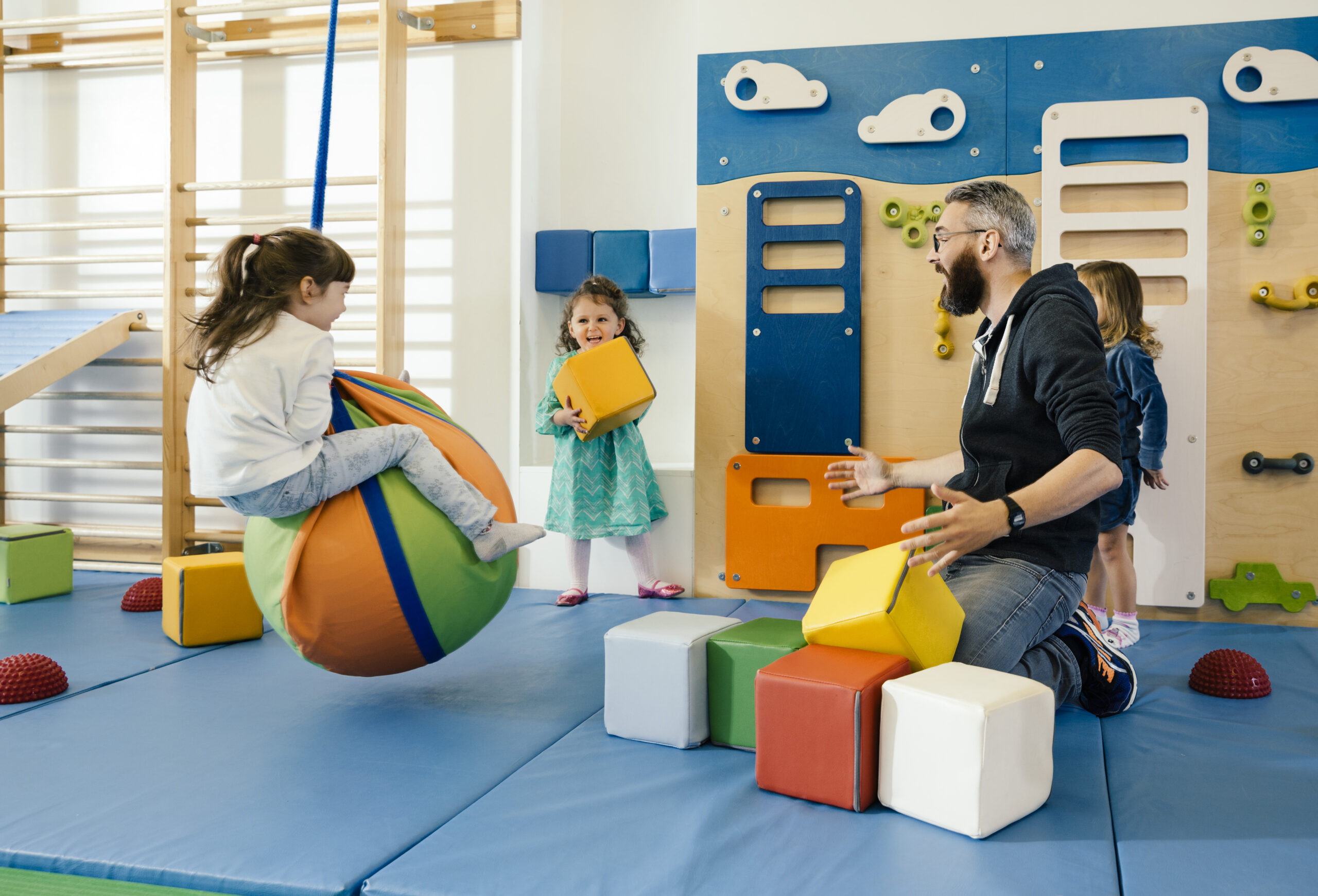 Ein männlicher Erzieher sitzt auf einer blauen Matte in einem hellen Turnraum und spielt mit drei Kindern. Ein Mädchen sitzt auf einem bunten Sitzball, ein anderes hält einen gelben Baustein hoch und ein drittes Kind steht daneben. Im Hintergrund sind eine Kletterwand und verschiedene Turngeräte zu sehen.