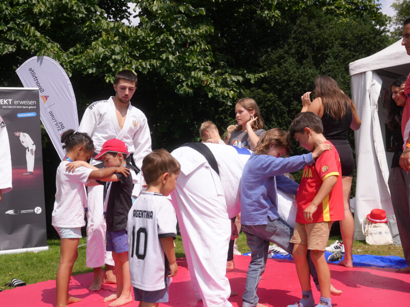 Eine Gruppe von Kindern und Jugendlichen übt gemeinsam Kampfsporttechniken. Ein Jugendlicher im weißen Judogi führt die Übungen vor, während ein Kind im argentinischen Trikot und ein anderes in roter Kleidung die Bewegungen nachahmen. Im Hintergrund sind weitere Kinder und Jugendliche zu sehen.