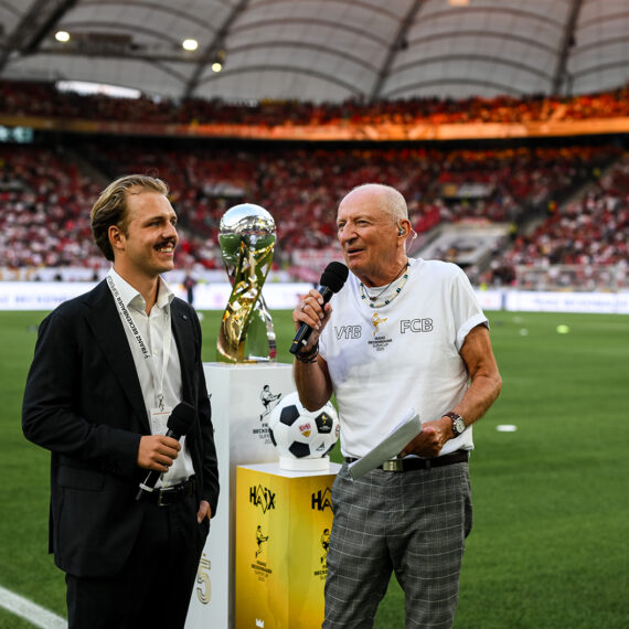 Joel Beckenbauer interviewt eine ältere Person vor dem Spiel des Franz-Beckenbauer-Supercups zwischen VfB Stuttgart und FC Bayern München im MHPArena Stadion. Im Hintergrund ist das Spielfeld zu sehen.