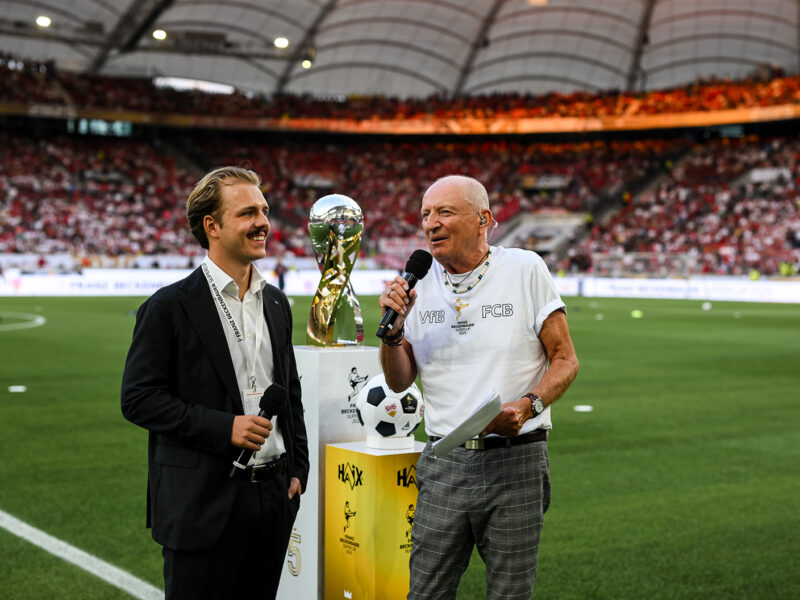 Joel Beckenbauer interviewt eine ältere Person vor dem Spiel des Franz-Beckenbauer-Supercups zwischen VfB Stuttgart und FC Bayern München im MHPArena Stadion. Im Hintergrund ist das Spielfeld zu sehen.