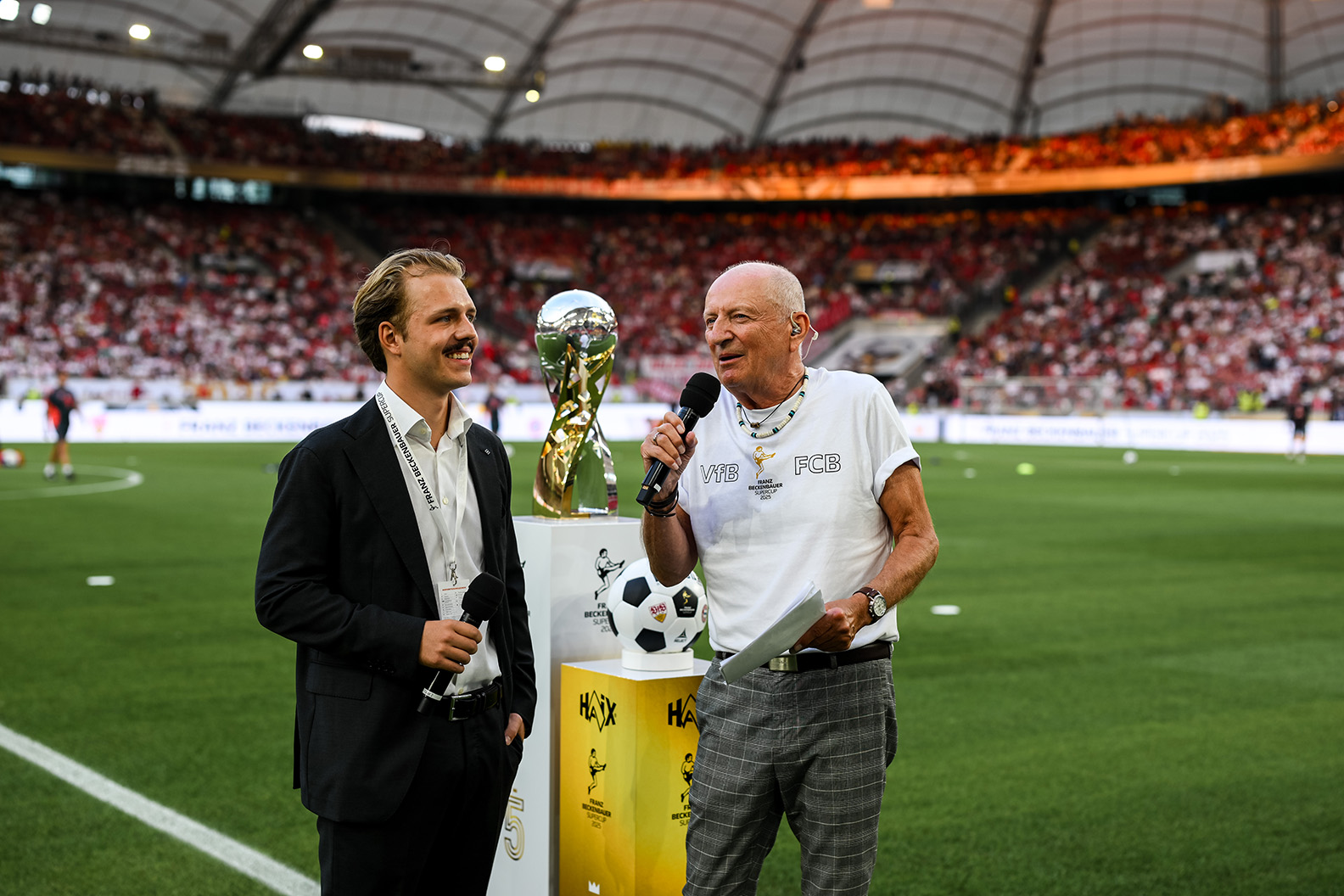 Joel Beckenbauer interviewt eine ältere Person vor dem Spiel des Franz-Beckenbauer-Supercups zwischen VfB Stuttgart und FC Bayern München im MHPArena Stadion. Im Hintergrund ist das Spielfeld zu sehen.