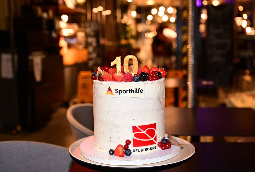 Festliche Geburtstagstorte mit der Aufschrift "10", dekoriert mit frischen Beeren und dem Logo der Deutschen Sporthilfe sowie dem Logo der DFL Stiftung. Die Torte steht auf einem silbernen Tablett vor verschwommenem Hintergrund.
